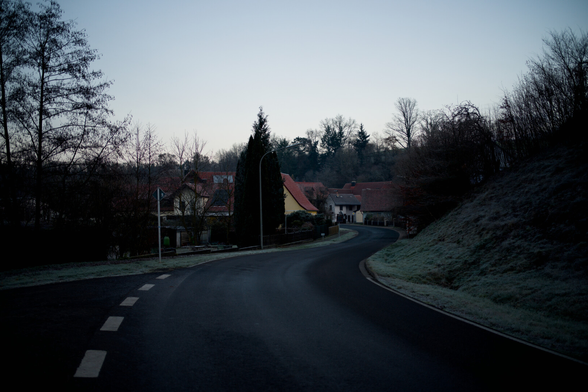 Die Straße Schlängelt sich durchs Bild. Rechts ein Ranga. (Hang) Mit Gestrüpp. Der in Häuser Übergeht in der Ferne. Links Gärten. Danach auch Häuser. Im Vordergrund geht eine Straße links ab. Überall vereinzelt Bäume.
