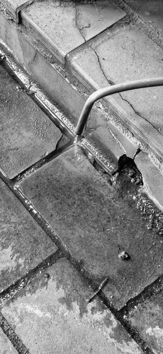 Schwarzweißfotografie eines im Durchmesser kleinen Röhrchens, das über einem Rinnstein abgeknickt ist und aus dem Wasser in den Rinnstein läuft. 