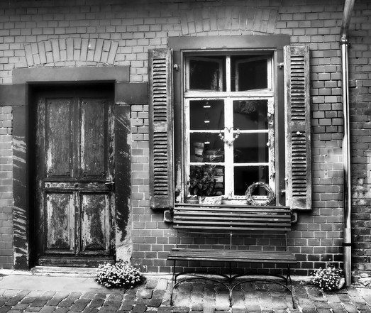 Auf dem Bild ist links eine alte Holztür und rechts davon ein altes Sprossenfenster. Aussen unter dem Sprossenfenster steht eine Holzbank. Ganz rechts sieht man das glänzende Fallrohr von oben kommend in den Boden gehend. Das Foto ist in Schwarzweiß.