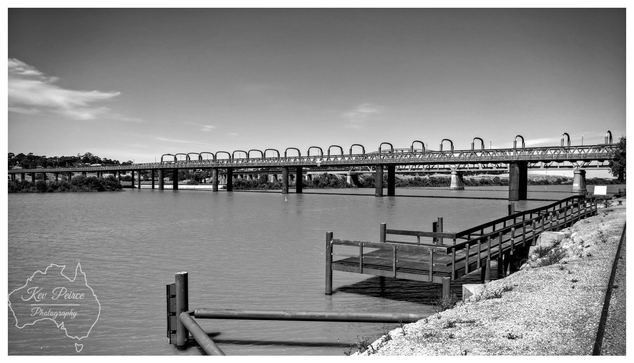 A black and white photograph of the historic Murray Bridge Rail Bridge spanning the wide, calm River Murray in South Australia.  The bridge, characterized by its distinctive repetitive arched ironwork, stretches across the centre frame.  In the foreground on the right, a wooden jetty and walking path extends over the gravel and bank beside the water. The sky is bright and clear. Photo by Kev Peirce.