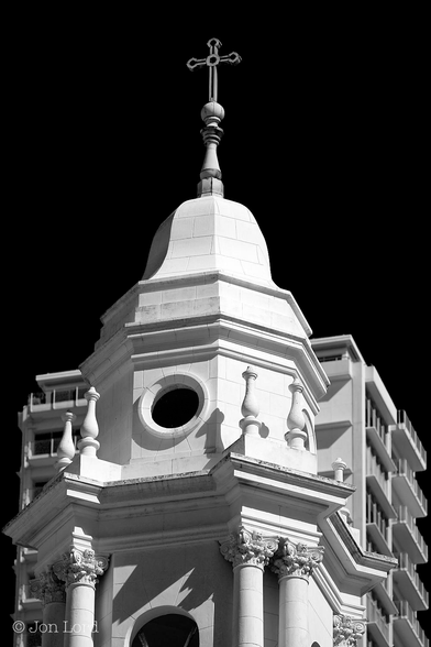 This is a high contrast black and white street / architectural photo of a church tower and high-rise apartment block in portrait format. San Francisco (2013).

In the foreground is the upper section of a brilliant white Spanish style church tower. The lower section in view has tall columns arranged in pairs in each of the four corners surrounding what looks like a ten sided white stone tower. Supported by the columns is an ornate plinth that supports the upper section which again appears to be ten sided, slightly tapered with a slightly flattened bell or cone shaped top - topped of course with a cross. 
immediately behind and partially obscured by the church tower is a square high-rise apartment building, also brilliant white, with balconies stretching across each floor. 
Surrounding each building is a clear and cloudless sky rendered jet black.
The church tower belongs to the former Nuestra Señora De Guadalupe Church, in Russian Hill, San Francisco.