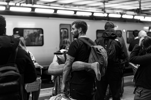 The image captures a candid moment in a train station, rendered in grayscale. The central focus is a man with a backpack, turned to his right, carrying a large, sculpted leg under his arm. The leg is a light color, contrasting with the man's dark clothing and the surroundings. He has a beard and visible arm tattoos.

Behind him, a train is visible, with its doors and windows partially obscured. Passengers are waiting, some with backpacks and coats. Their faces and expressions are mostly hidden, emphasizing the sense of anonymity of a public space.
