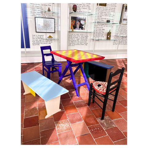 A colorful table with a red and yellow checkerboard design is surrounded by mismatched chairs, including a blue chair, a black chair with a decorative seat, and a simple light blue bench. The background features a wall with glass displays and handwritten text.