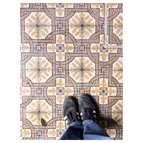The image features a close-up view of a patterned floor made of colorful tiles, showcasing intricate designs with floral motifs in various shades. A pair of black sneakers stands on the floor, partially visible at the bottom of the image.