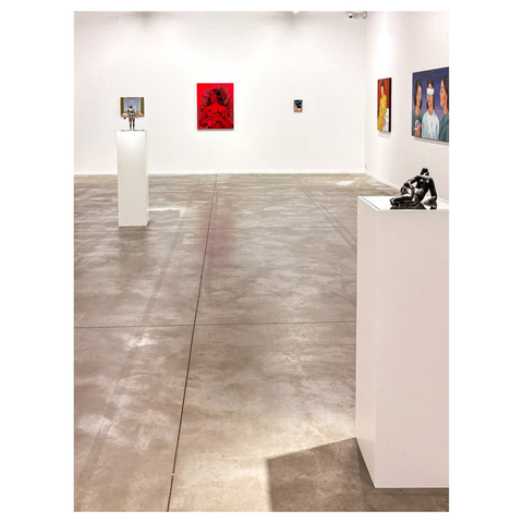 An art gallery interior featuring white walls and a polished concrete floor. Two sculptures on white pedestals are present, along with several colorful paintings displayed on the walls. The artworks include a prominent red piece. The space is well-lit and minimalistic.