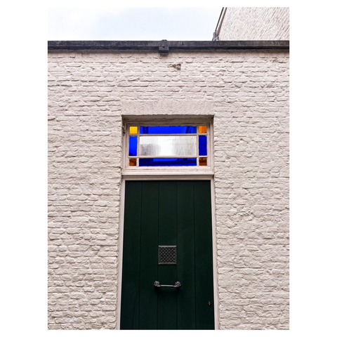 A green door with a decorative handle and a metal vent is set against a textured white brick wall. Above the door, there is a rectangular stained glass window featuring blue, orange, and yellow glass sections.