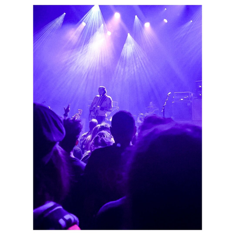A live music performance by High On Fire featuring guitarist Matt Pike on stage with purple lighting. The audience is visible in the foreground, raising their hands in excitement.