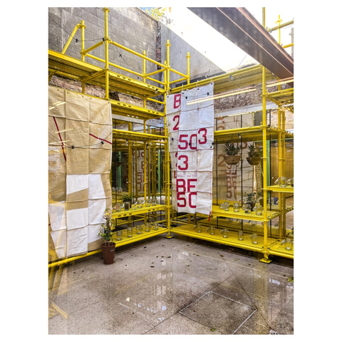 An outdoor space seen from inside, featuring construction scaffolding painted yellow. The area displays large fabric banners with red text and contains shelves with glass objects. A small potted plant is visible on the floor, and the surface appears wet from recent rain.