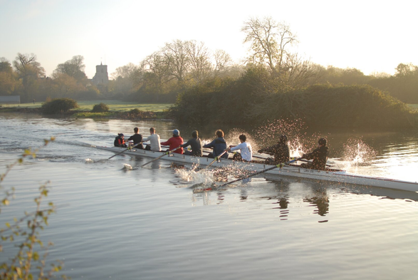 A rowing eight on a river with a village church in the background. One of the rowers just caught a crab and there's a big spray of water