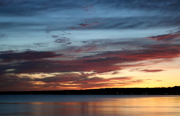 Photograph of a colourful sunrise over a bay, with the coastline on the horizon. The sky is glowing red near the horizon to the left and mostly yellow to the right, with pink reflected off horizontal clouds in the middle of the sky, and blue at the very top. The colours are reflected on the almost calm sea below. A few house lights illuminate the coastline, which appears black.

Photographie d'un lever de soleil coloré au-dessus d'une baie, avec la côte à l'horizon. Le ciel est rougeoyant près de l'horizon vers la gauche et plutôt jaune à droite, avec du rose qui se reflète sur les nuages horizontaux au milieu du ciel, et du bleu complètement en haut. Les couleurs se reflètent sur l'eau de la mer presque calme dans le bas. Quelques lumières de maisons illuminent la côte qui apparaît noire.