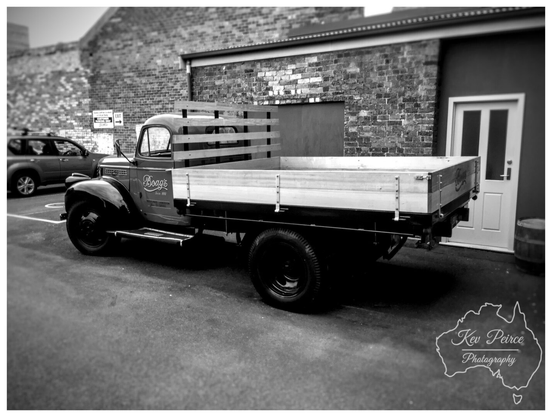 Black and white photograph of a beautifully maintained, vintage Boag's brewery flatbed truck, signed by Kev Peirce Photography.  The truck is parked in an urban setting, possibly near the brewery itself, against a backdrop of dark brick and concrete buildings.  The truck features low, wooden slatted sides on the bed and 'Boag's Since 1881' written on the side of the cab.  Its classic body lines and dark wheels stand out, creating a strong contrast with the bright white trim of the nearby building and the light paint of the truck bed.
