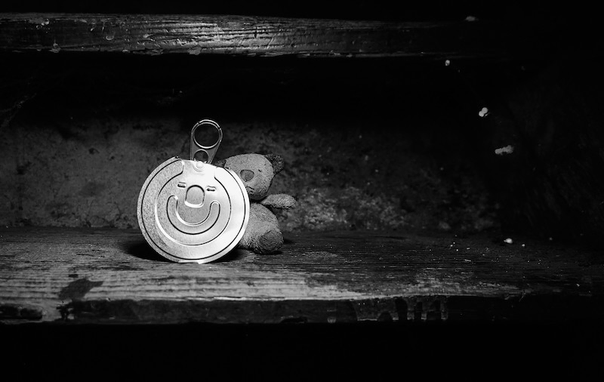 Man sieht einen traurigen kleinen Teddybär teilweise hinter einem Deckel von einer Erdnussdose hervorschauen. Der Deckel ist aus glänzendem Metall gefertigt und oben sieht man die Lasche mit der man den Deckel von der Erdnussdose öffnet. Auf dem Deckel selbst sind Einstanzungen die wie ein lächelndes Gesicht aussehen. Das Foto ist in Schwarzweiß.