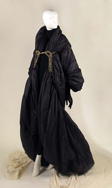 an image of a mannequin wearing a long black jacket / dress garment, gold chains across the chest of it, the jacket goes down to the floor, the mannequin is seen from the front in a three quarter view 