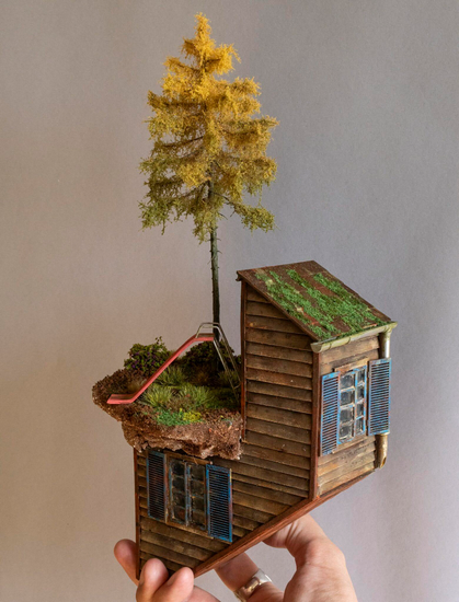 a hand-sculpted miniature by Simon Laveuve, held by the artist's hand for scale, of a shack-like building with a tree and a children's slide