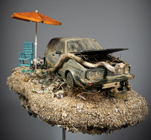 a hand-sculpted miniature by Simon Laveuve of an abandoned car with a large root growing through the hood, with a beach umbrella propped up next to it