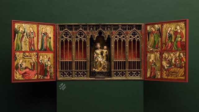 On a box pedestal against a wall, both painted deep green, an intricately carved and painted altarpiece in the form of a box with two doors opening to the side. The inside of the doors are brilliantly painted and gilded with scenes of people, angels, and beasts. The interior of the box contains a Madonna and Child, a statue fully in the round, gilded; framed by a carved "altar screen" device of gothic arches and traceries, gilded and painted. The whole is amazingly well preserved for something almost 700 years old.