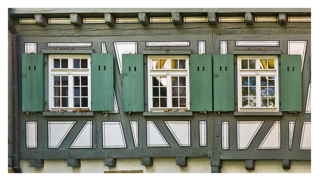 This is a detailed close-up photograph of the upper part of a traditional German half-timbered house. The wall features the characteristic exposed dark wooden beams forming a geometric pattern of rectangles, diagonals, and triangles, with the spaces between them filled with white plaster.There are three six-pane windows in a row at eye level. Each window has white frames and is surrounded by green wooden shutters that are currently open and fastened flat against the wall on both sides. The shutters are a muted sage green color with simple rectangular cut-outs near the top.Above and below the windows runs a horizontal row of closely spaced, protruding wooden beam ends. The timber framing is painted dark gray-brown, and the overall appearance is well-maintained and typical of historic buildings found in many German towns.