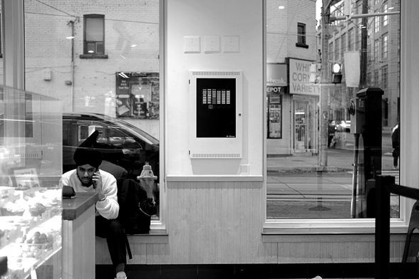 In a corner coffee shop. There are two windows in the background, lit by early twilight. The one on the right shows the convenience store across the street. In the lower corner of the image, on the ledge of the left window, a man in a turban sits, talking on his phone. A car passes behind him.