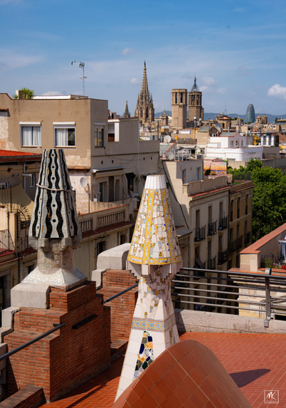 Color photo from a rooftop with sculptural chimneys looking out across the city of Barcelona. 