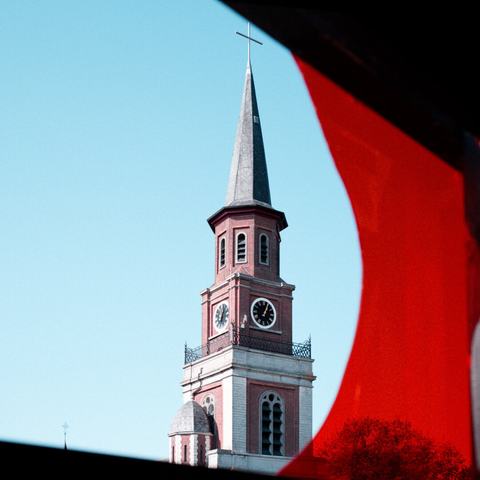 The spire of a church, with cross on top, against a blue sky background. The image is framed by a bright-red piece of translucent plastic along the right-hand side.