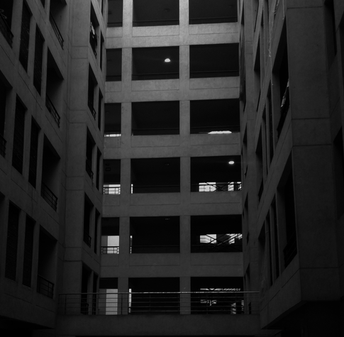 Dark grayscale image of a multi-story building interior, featuring vertical rows of windows and railings. The atmosphere is gloomy and minimalistic.