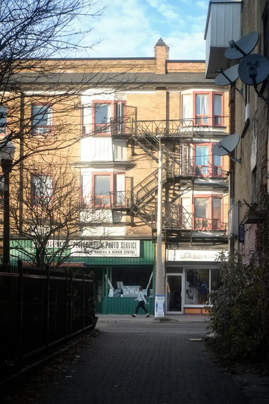 Standing along a small alley looking out towards a city street a pedestrian can be seen walking along the far sidewalk. Tall buildings with different sized windows and shops can be seen, a fence lines the alley on the left and a tall building with satellite receivers can be seen on the right.
