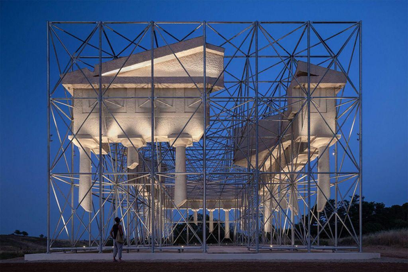 A person stands beneath an artistic structure resembling ancient architecture, illuminated against a twilight sky, supported by metal scaffolding.