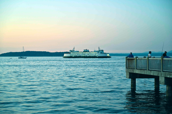 A ferry approaching (or departing from) a terminal just out of frame. It's dusk on a clear night, and the sky is a gradient from pale blue through yellow to pale pink. On the right side of the screen, there's a pier.