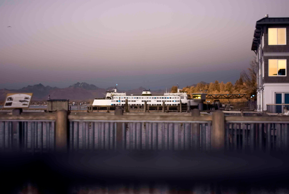 A ferry at a dock, with a pier and a railing crossing the bottom of the frame. The vegetation looks yellow and brown now, and the image generally has a grey colour cast to it. Lights that would normally be green or even white are bright blue.