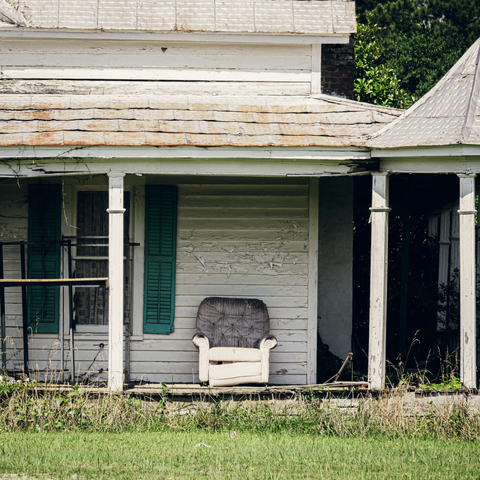 An old white wooden house with severely deteriorated exterior showing extensive paint peeling and weather damage. The front porch features white square columns supporting a covered overhang with worn green trim. A single weathered upholstered armchair with a patterned back sits centered on the porch. Dark doorways and windows frame the chair, with teal shutters visible on the left window. The yard shows overgrown grass and weeds in the foreground, while dense green foliage and trees are visible in the background. A portion of the shingled roof is visible at the top of the frame, showing signs of age and wear.