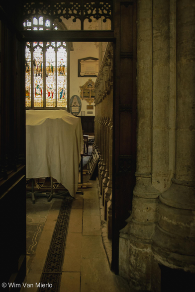 Church interior: looking through an opening in a wooden screen (next to a stone pillar) towards a stained-glass window; some furniture is covered in white sheets to protect it from grime.