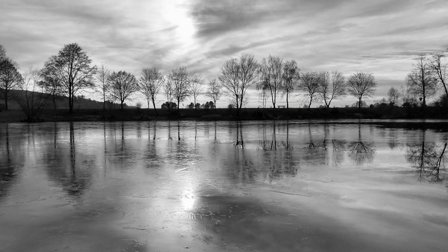 Man sieht auf einen See, auf dessen Oberfläche sich teilweise schon Eis gebildet hat. Am anderen Ufer stehen Bäume ohne Blätter die sich auch auf der leicht vereisten Oberfläche vom See spiegeln. Am Himmel sieht man einige Schleierwolken. Das Foto ist in Schwarzweiß.