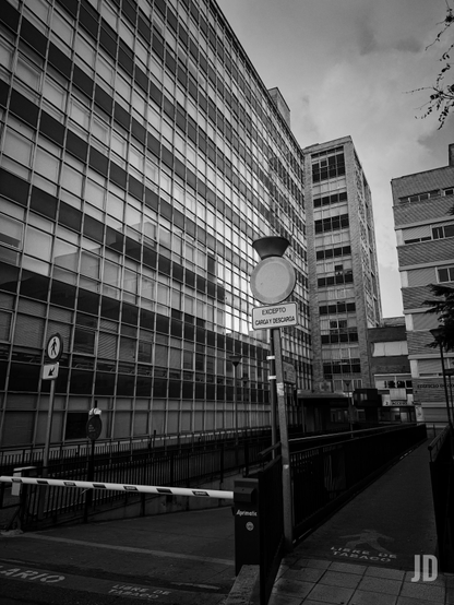 La imagen es una fotografía en blanco y negro que muestra una vista urbana de edificios de oficinas altos.
• Primer plano: Domina una gran estructura moderna de cristal y hormigón con numerosas ventanas rectangulares. A su derecha, en un pequeño paso peatonal/acceso, hay una señal de tráfico circular con la inscripción "EXCEPTO CARGA Y DESCARGA".
• Fondo: Se ven otros edificios de altura similar, con fachadas también cuadriculadas. La luz sugiere un día nublado o una iluminación tenue, acentuando el contraste dramático del blanco y negro.
• Zona baja: Se aprecia una rampa de acceso o entrada con una barrera blanca y negra y lo que parece ser una zona de acera y asfalto con texto en el suelo ("LIBRE DE TABACO"). 