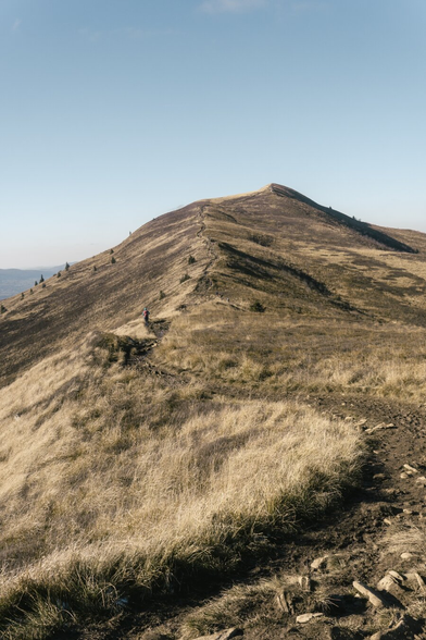 Mountain hills covered with dry grass, grass and rocks in the foreground, and a trail leading to the summit in the background.