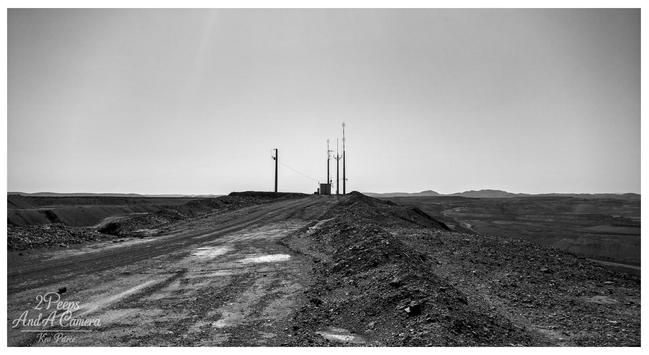 A low angle, black and white photograph signed by Kev Peirce. A rough, dirt track littered with rubble leads up a steep embankment toward a cluster of utility poles and a radio communication tower in the distance.

The foreground is heavily textured with shadows and loose rock. The horizon is wide and flat, with low hills visible in the far background under a bright, clear sky.

The landscape suggests an arid, remote, or mining affected area, likely near Leigh Creek.
