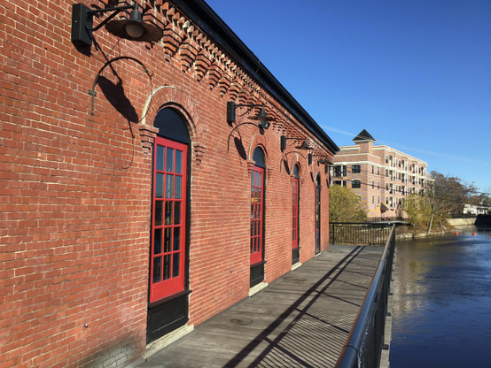 Brightly sun-lit red-painted windows in a brick-walled tavern in Nashua, New Hampshire, with a walkway along a canal (or river?) looking at other buildings in the distance against a clear blue sky.