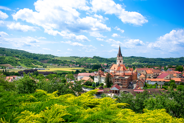 A photograph of a town, blue sky with clouds, hills and greenery can be seen around it.