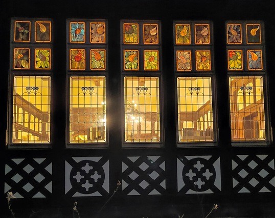 Durch 5 hohe Fenster kann man in das beleuchtete Innere schauen. Über den 5 Sprossenfenstern sieht man zusätzlich noch je 4 kleinere Fenstereinheit mit Tiffany. Diese zeigen bunte Blumen und leuchten durch das Licht im Inneren. Unter den Fenstern erkennt man in dem dämmrigen Bild noch die verzierte Fachwerkfassade.