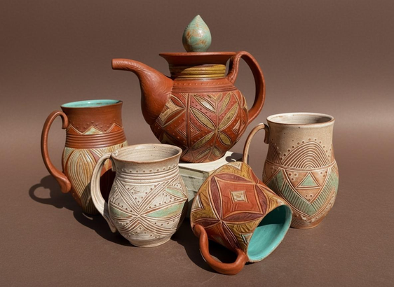 A collection of intricately designed ceramic pottery, including a teapot and several mugs, showcasing various warm colors and geometric patterns. The pottery is arranged on a neutral brown background. 