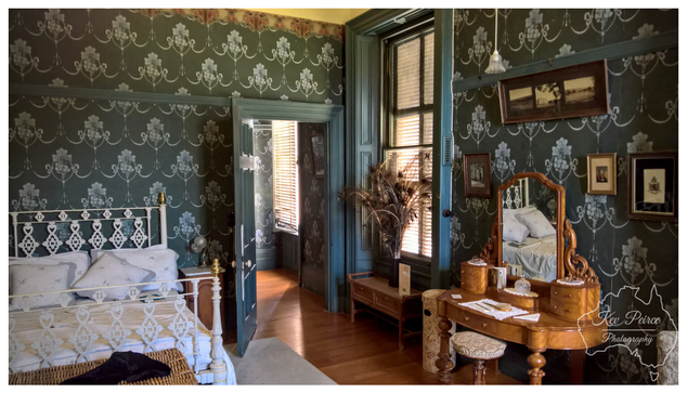 A well preserved Victorian era bedroom at Martindale Hall. The room features dark teal and white patterned wallpaper, polished wooden floorboards, and tall windows with wooden blinds casting long shadows.

On the left is a white wrought iron bed with white linens. To the right, a rich wooden dressing table with an ornate mirror holds various grooming items and reflects the bed.

The door in the centre is open, leading to another richly decorated room bathed in natural light. The photograph is signed Kev Peirce.