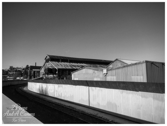 Black and white photograph of the Launceston Old Railway Workshops. The foreground is dominated by a sharply curving railway track running parallel to a long, corrugated metal fence that reflects the bright light.

Behind the fence, several large, old railway maintenance sheds and industrial buildings with corrugated iron roofs rise up against a clear, bright sky.

