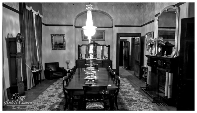 A wide, black and white photograph of the formal dining room at Martindale Hall. A long, dark wooden dining table dominates the center of the room, set with decorative plates and surrounded by matching chairs.

Above the table hangs a large, sparkling crystal chandelier, which is brightly lit and forms the central focal point under a high arched ceiling.

To the right is an elaborate fireplace and a tall, arched mirror reflecting the room. On the left stands a grandfather clock and heavy drapes.

The monochrome treatment emphasises the room's historical elegance and drama. The photograph is signed Kev Peirce.