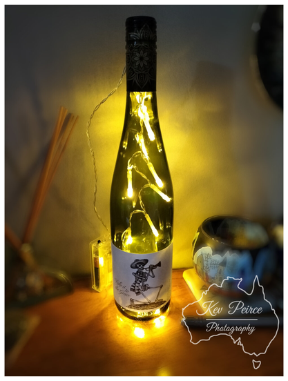 Color close up photograph of a decorative green glass wine bottle repurposed as a lamp, signed by Kev Peirce Photography.  A string of warm, yellow fairy lights has been inserted into the bottle, illuminating the glass and the surroundings. A white label on the bottle features an illustration of a festive skeleton playing a trumpet.  The bottle sits on a wooden surface next to a glass diffuser with reeds and a small, decorated ceramic bowl, bathed in the soft glow of the lights.
