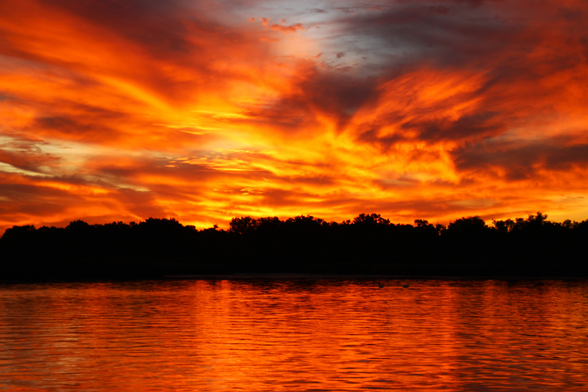Photograph of a spectacular sunset with bright yellow and orange clouds seeming to explode over a lake that reflects the colors.