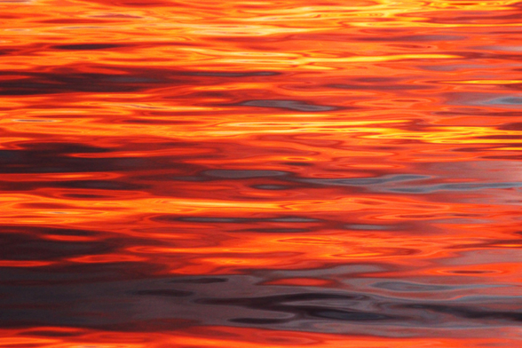 Photograph of sunset reflections of yellow, orange, red, and violet light forming horizontal swirls on the surface of a lake.