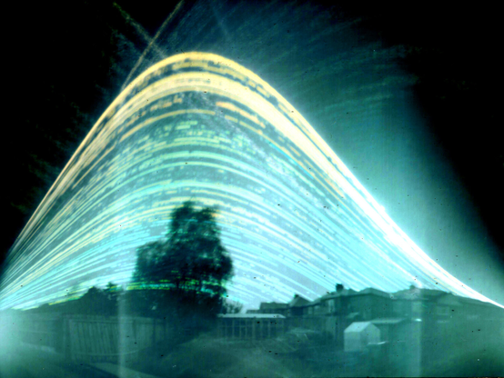 A series of parabola-shaped light lines, from the sun's highest point to its lowest. A tree and houses stand as ghosts in the foreground.