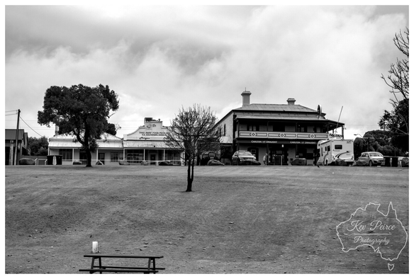 A black and white photograph by Kev Peirce shows the historic streetscape of Morgan, South Australia.  The image features a wide, dusty foreground leading to two classic Australian buildings: a single story shop on the left and a two story pub, the Morgan Wharf Hotel (formerly the Commercial Hotel), on the right.  A prominent, nearly bare tree stands in the centre foreground, and a picnic bench is visible near the bottom edge. Cars and a caravan are parked outside the pub under a cloudy sky.