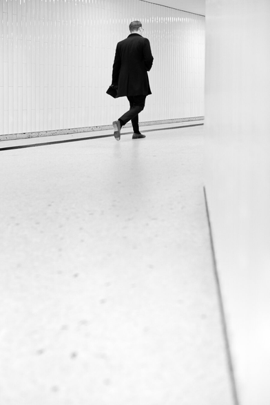 A man in a black business suit rushes through the corridors of a subway station, which is designed entirely in white, on his way home.