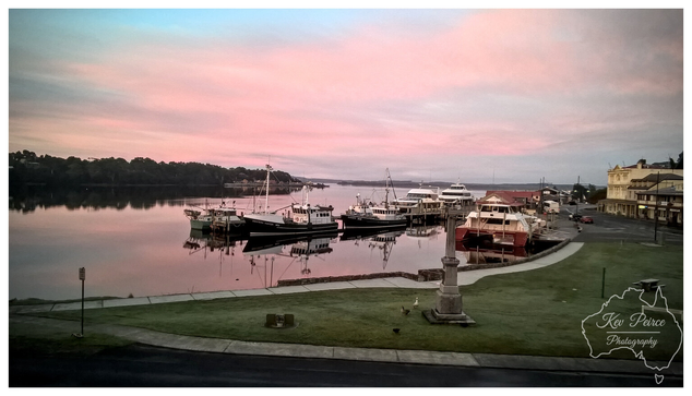 A tranquil, wide angle photograph of the historic Strahan waterfront at sunset. Fishing boats and larger vessels are moored at a jetty reflected in the calm, smooth waters of the harbour.  The sky above is dominated by soft, expansive pink and gold hues. A grassy park and a commemorative stone monument are visible in the foreground. Photo by Kev Peirce.