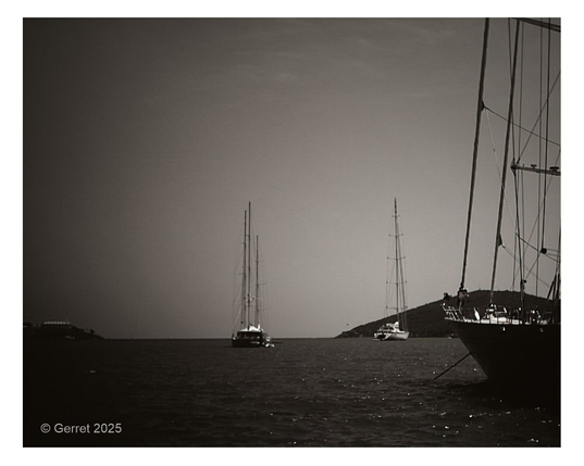 Black and white photo of sailboats anchored in calm waters. Two boats are centered with tall masts, while another is silhouetted on the right. A peaceful, nostalgic tone.